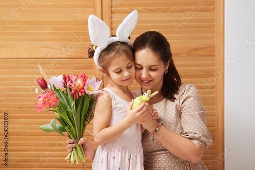 Young beautiful mother and her little daughter in the dress with white rabbit's ears on her head hold flowers and dyed eggs in their hands on the background of wooden doors