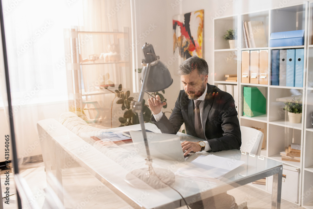 Serious handsome bearded office manager in suit sitting at table and checking online files on laptop while working on report Serious handsome bearded office manager in suit sitting at table and checking online files on laptop while working on report