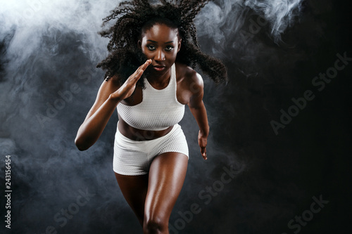 Fototapeta Naklejka Na Ścianę i Meble -  Strong athletic black skin woman sprinter, running on background with smoke wearing in the sportswear. Fitness and sport motivation. Runner concept with copy space.