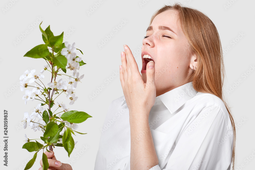 Isolated shot of young cute woman being allergetic to spring blossom, sneezes, keeps mouth widely opened, poses against white background, doesnt like odour. People, sickness and allergy concept Isolated shot of young cute woman being allergetic to spring blossom, sneezes, keeps mouth widely opened, poses against white background, doesnt like odour. People, sickness and allergy concept