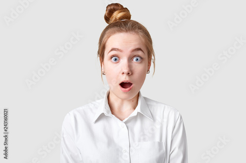 Image of surprised teenage girl has bugged blue eyes, keeps mouth widely opened, dressed in elegant shirt, isolated over white background, being in stupor. People, reaction, shock, surprisement