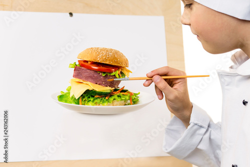 cute boy in chef uniform preparing with a brush on paper an appetizing sandwich on a white background