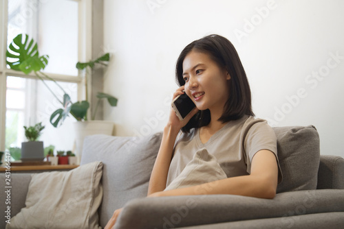 Happy young Asian woman using mobile phone talking with her friend with smile while sitting on a couch at home in the living room. Technology and lifestyle concept.