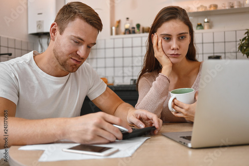 Casually dressed young couple managing finances at home: bearded husband calculating expenses using calculator while his wife with mug paying domestic bills online on generic portable computer