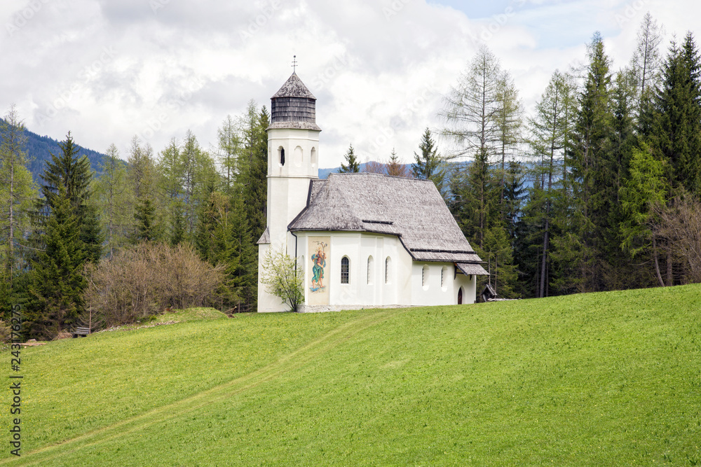 View of a small white church on a hill in the middle of a vibrant green alpine meadow in Ehrwald, Austria.