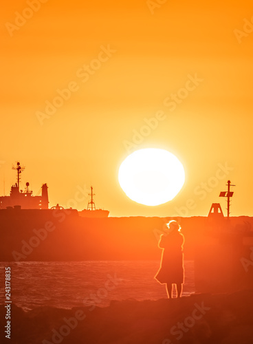 Fuzzy silhouette of woman taking picture of red sunset in a harbor.