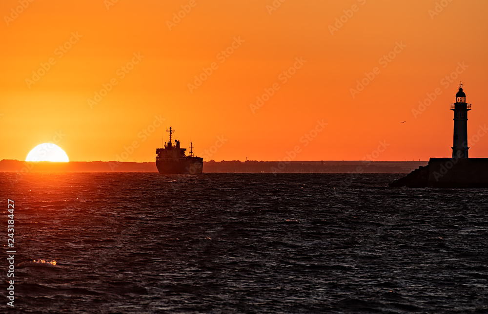 Hazy ocean sunset with ship and lighthouse silhouettes.