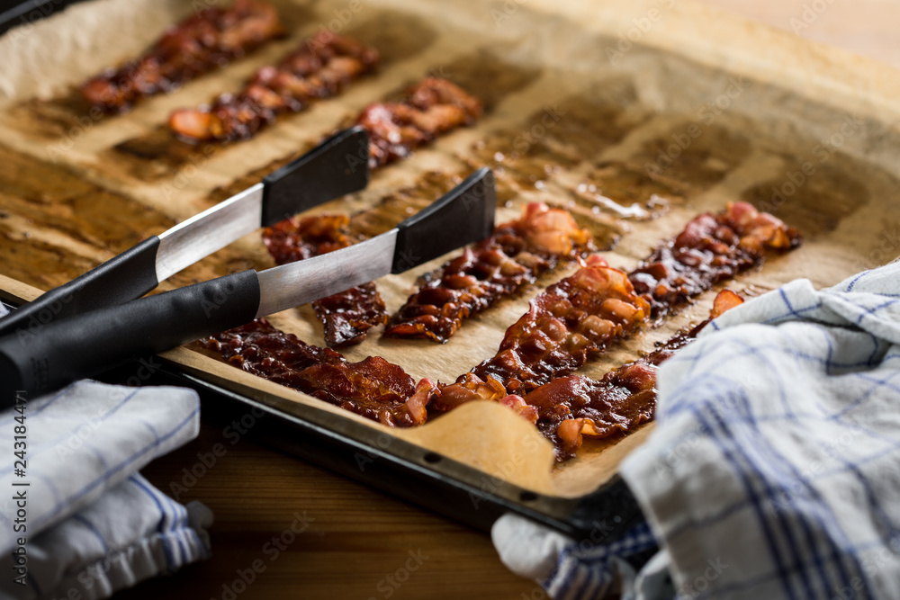 Knusprig gebratene Bacon Speck Scheiben aus dem Ofen auf Backblech mit Backpapier mit Geschirrtuch und Zange in Küche Knusprig gebratene Bacon Speck Scheiben aus dem Ofen auf Backblech mit Backpapier mit Geschirrtuch und Zange in Küche