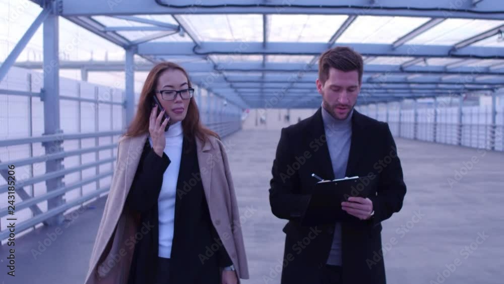 Businessman writing notes on a tablet and asian businesswoman talkingby phone walk together. Businessman writing notes on a tablet and asian businesswoman talkingby phone walk together.