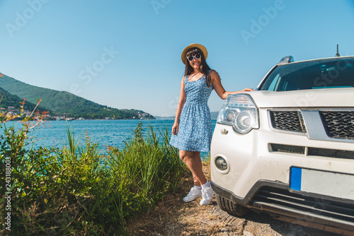 woman in blue dress standing near white car at seaside with beautiful view on bay and mountains © phpetrunina14