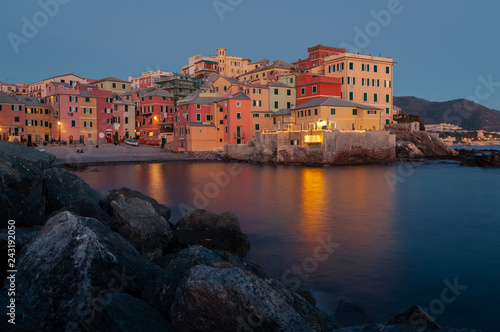 Genoa, Italy. Ancient village of Boccadasse during the blue hour. On the right a small beach in the middle of old pastel-colored houses. Reflections of the lights on the sea. Rocks in the foreground. 