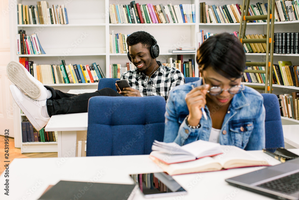 Happy afro-american student at university library sitting and laying legs on the table, listening to music and working with a smartphone. An African American girl prepares for exams in the library Happy afro-american student at university library sitting and laying legs on the table, listening to music and working with a smartphone. An African American girl prepares for exams in the library