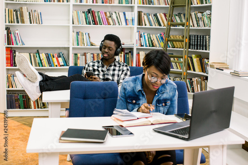 Afro Americans students at the library. The girl works in a library using a laptop and reading books on the background of a guy who listens to music and put his legs on the table.