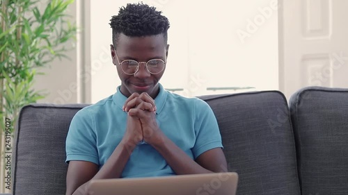 Smiling, young man using laptop at home. Handsome, african american man in casual blue t-shirt and classic eyeglases rejoice succes. Freelancer succes concept. photo