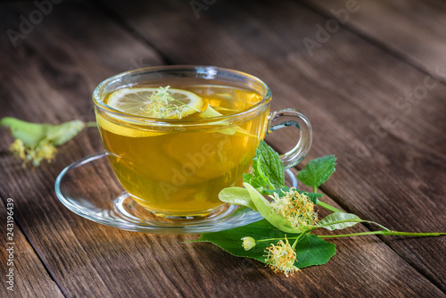 Cup of lime tea, standing on a wooden table, surrounded by fragrant linden flowers, in the rays of sunlight.