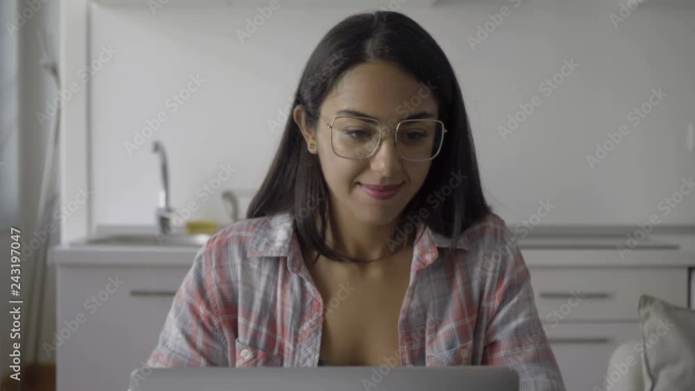Smiling brunette woman using laptop at kitchen. Young girl in eyeglasses typing on laptop at home. Technology concept Smiling brunette woman using laptop at kitchen. Young girl in eyeglasses typing on laptop at home. Technology concept