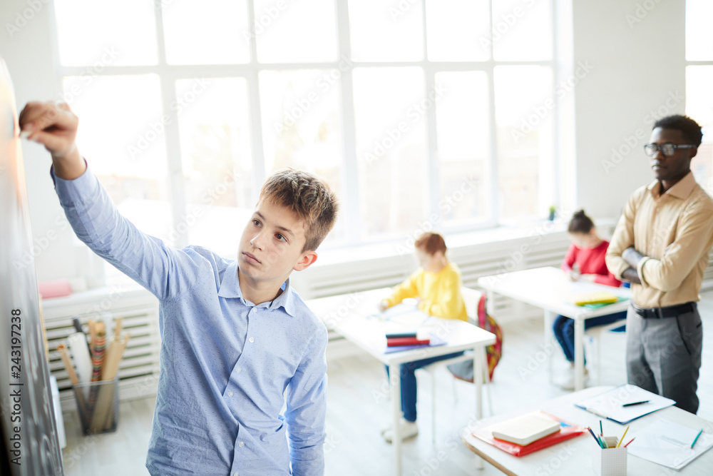 Serious schoolboy writing something on blackboard at lesson in front of teacher and classmates