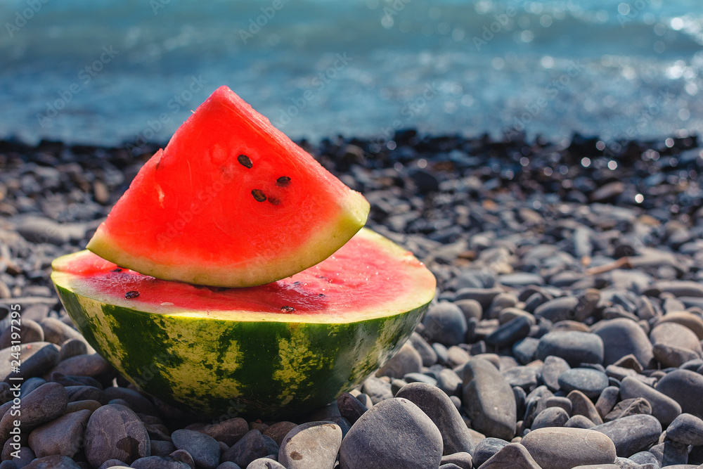 A large, juicy, cut watermelon, lies on a pebble beach on the shore of the blue sea. A large, juicy, cut watermelon, lies on a pebble beach on the shore of the blue sea.
