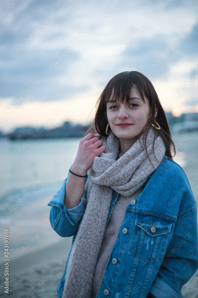 young pretty girl standing in jeans jacket with warm scarf on the beach near the sea in the winter day