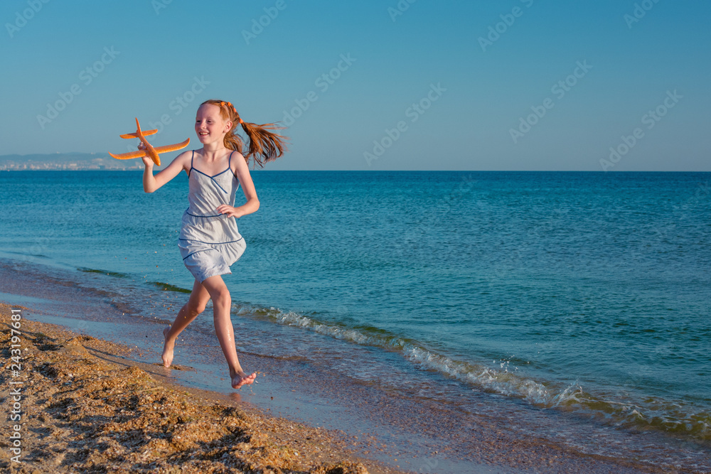 Red-haired girl, playing with a toy airplane, runs along the beach of the sea against the blue summer sky and enjoys the rays of the sun and freedom..