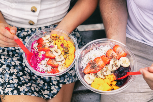 Acai bowl and pitaya dragonfruit smoothie healthy breakfast bowls young friends eating together. Couple man and woman eating sitting outside in park for lunch break. Closeup on food.