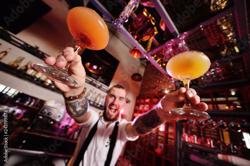 handsome bartender prepares an orange alcoholic cocktail and smiles on the background of a bar or a night club