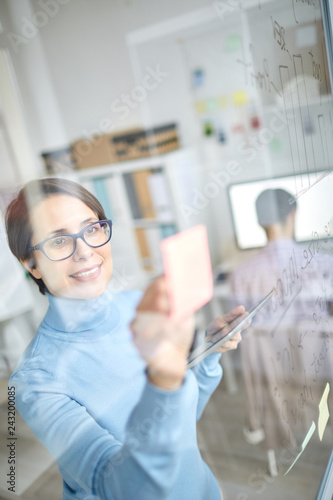 Pretty casual businesswoman standing by transparent board and reading workflow notes in office