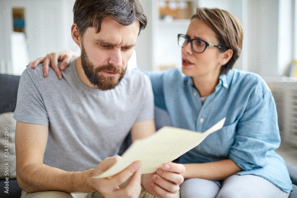 Confused man reading paper with answer to his application for job vacancy while his wife trying to reassure him Confused man reading paper with answer to his application for job vacancy while his wife trying to reassure him