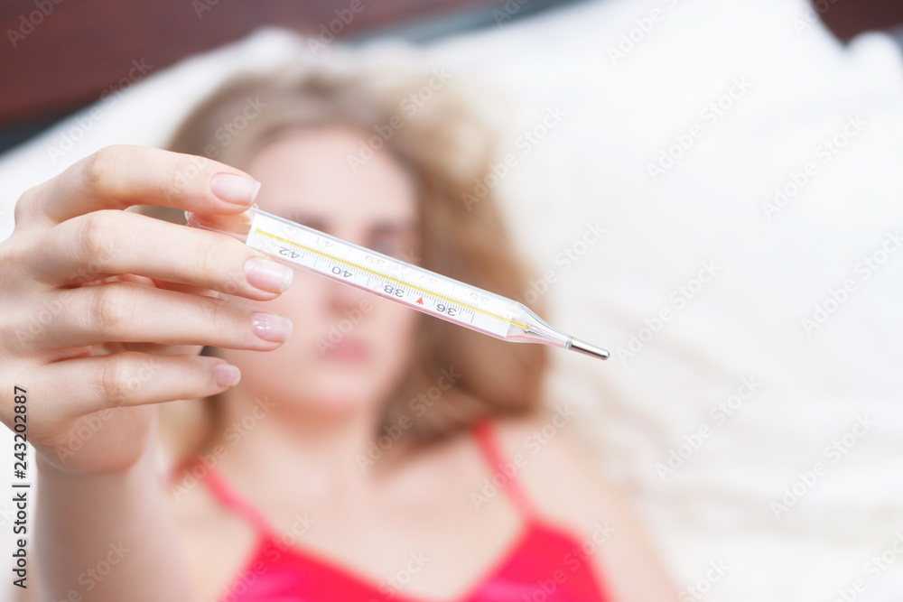 Woman with flu virus lying in bed, she is measuring her temperature with a thermometer Woman with flu virus lying in bed, she is measuring her temperature with a thermometer