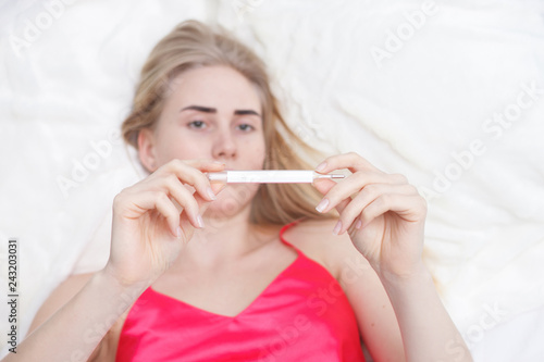 Young woman sitting on a couch, having a fever, holding a thermometer after measuring temperature