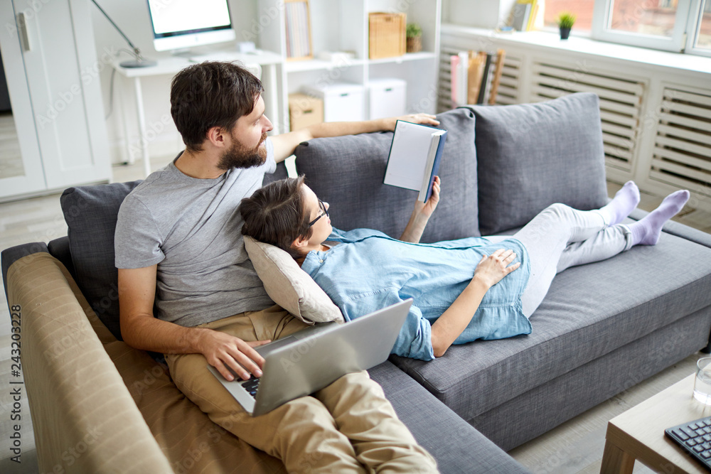 Young couple reading book or notes in notebook while relaxing on sofa in living-room on weekend