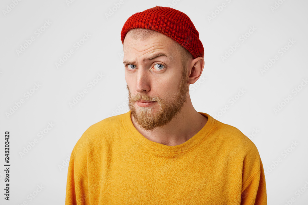 Portrait of bearded guy looks into camera suspiciously interrogatively with curiosity as if he is peeping, wearing a red hat and sweater isolated against a white background. Man is prying a keyhole. Portrait of bearded guy looks into camera suspiciously interrogatively with curiosity as if he is peeping, wearing a red hat and sweater isolated against a white background. Man is prying a keyhole.