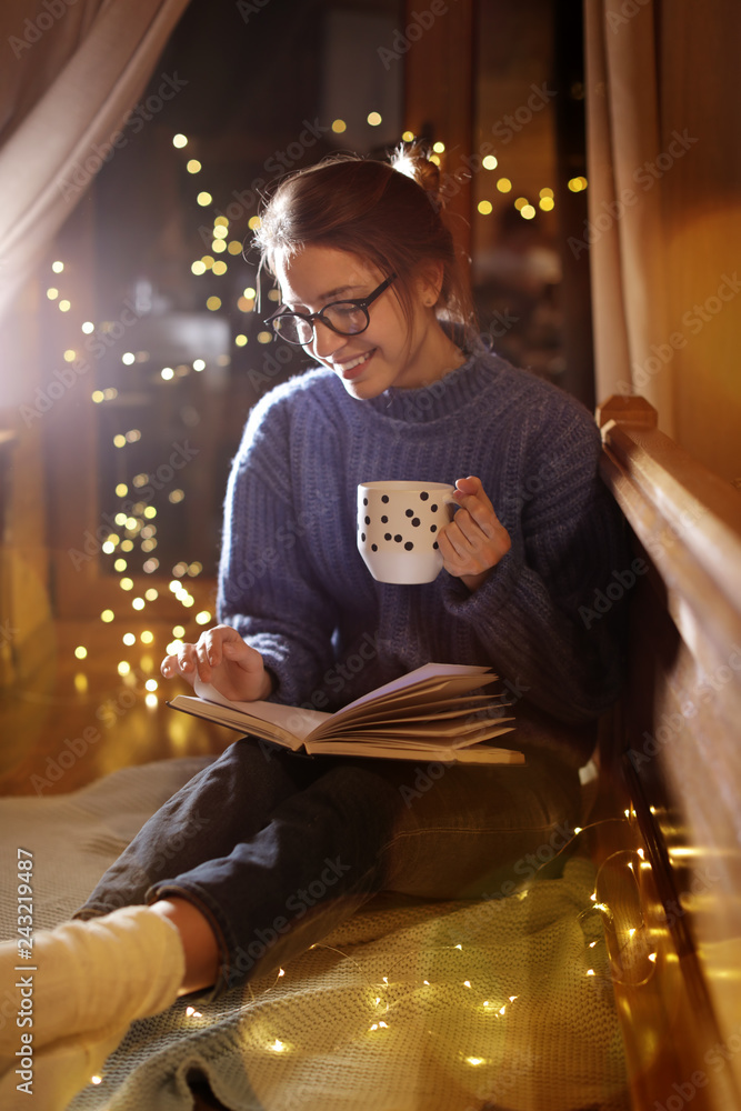 Woman with cup of hot beverage reading book at home in winter evening