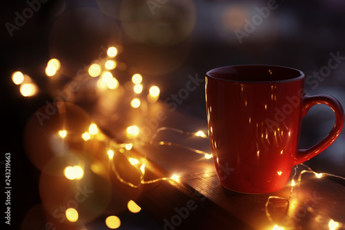 Cup of hot beverage on balcony railing decorated with Christmas lights, space for text. Winter evening
