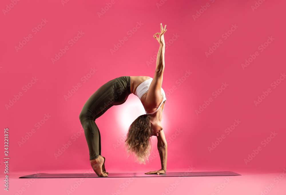 Beautiful advance yogi showing her incredible flexibility on her yoga mat in studio against a bright pink background with a light flare behind her. 
