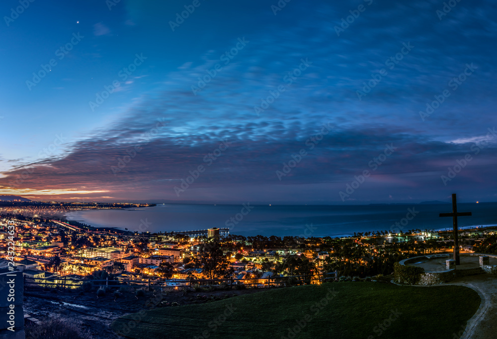Moon and Venus shine along with city lights of Ventura along the Pacific Ocean coast as dawn begins to light the cloudy sky.