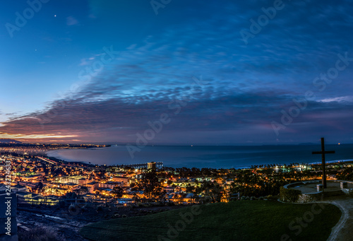 Moon and Venus shine along with city lights of Ventura along the Pacific Ocean coast as dawn begins to light the cloudy sky.