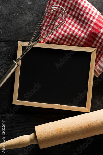Blank, empty, black chalkboard with wooden rolling pin, wire whisk and red checkered dish towel flat lay from above on black wooden table