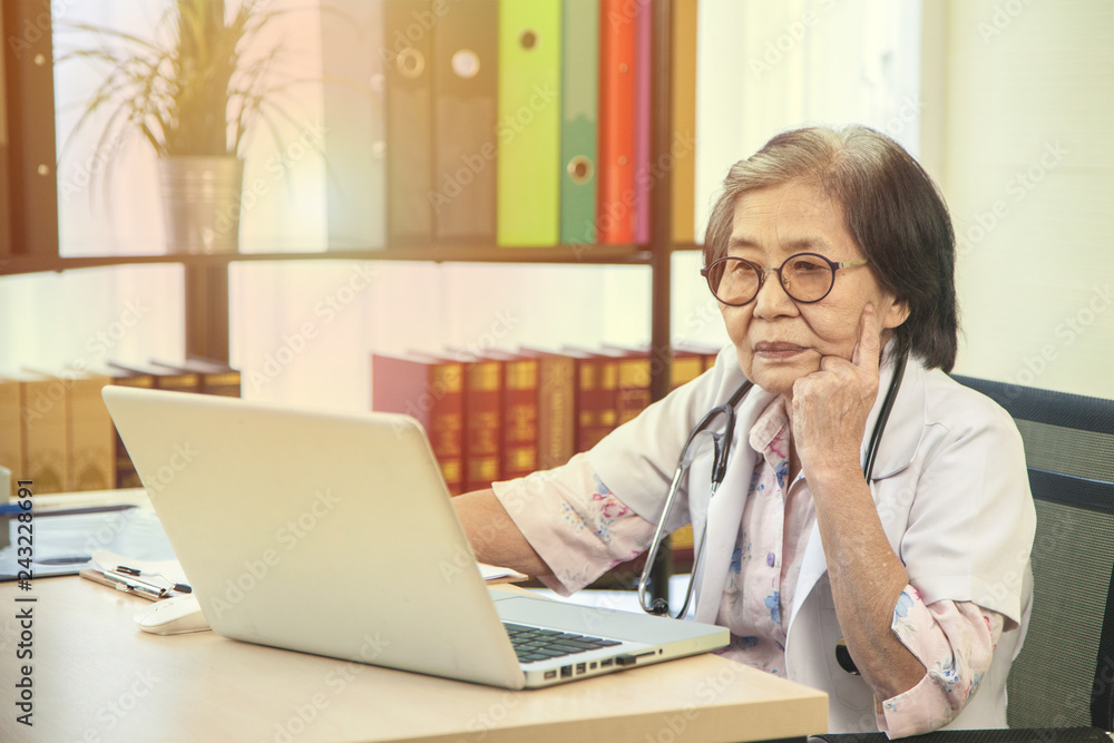 Old woman doctor sit and look at her laptop. Her smile for the health of patient in hospital - health care concept - Image Old woman doctor sit and look at her laptop. Her smile for the health of patient in hospital - health care concept - Image