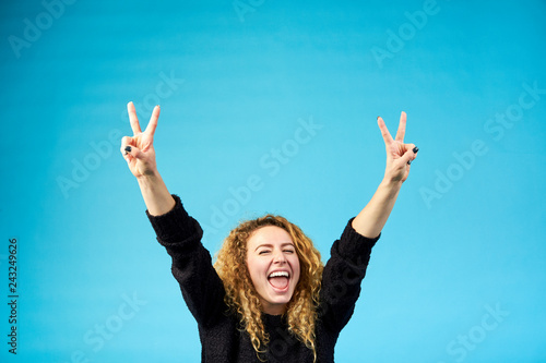Emotional satisfied young attractive ginger curly woman with opened mouth celebrating and cheering a success raising up hands with the sign of victory on blue background.