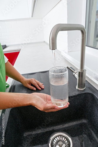 The girl fills a glass with water from a water tap in the kitchen.