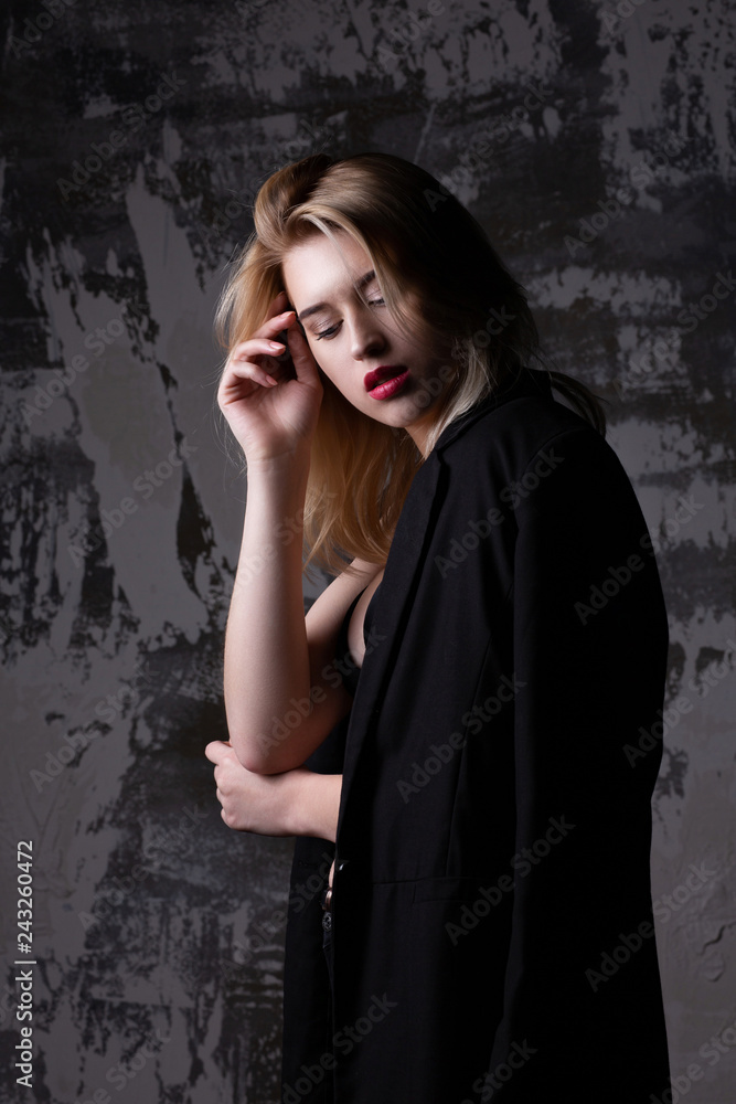 Tender blonde model wearing black jacket posing at studio with shadows