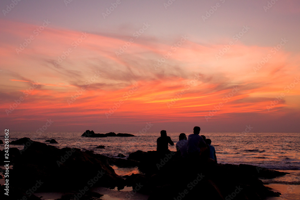 black silhouettes of friends sitting on stones against the ocean and purple pink red blue sunset sky