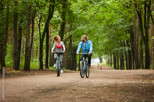 Young active man and woman sitting on bicycles while moving along road in park and talking to one another