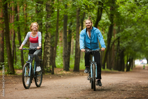 Happy guy and his girlfriend on bicycles looking at you while riding down forest road or in park