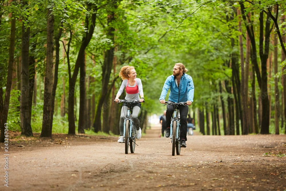 Young man and woman in activewear talking while cycling on road in park on summer weekend