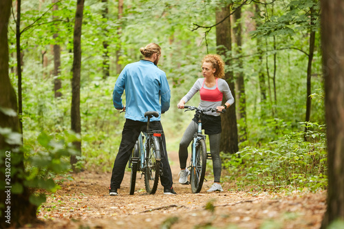 Active girl and guy with bicycles standing on forest path and discussing what way to choose for ride