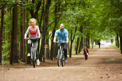 Two young active cyclists riding on bicycles along wide road between trees in park on summer day