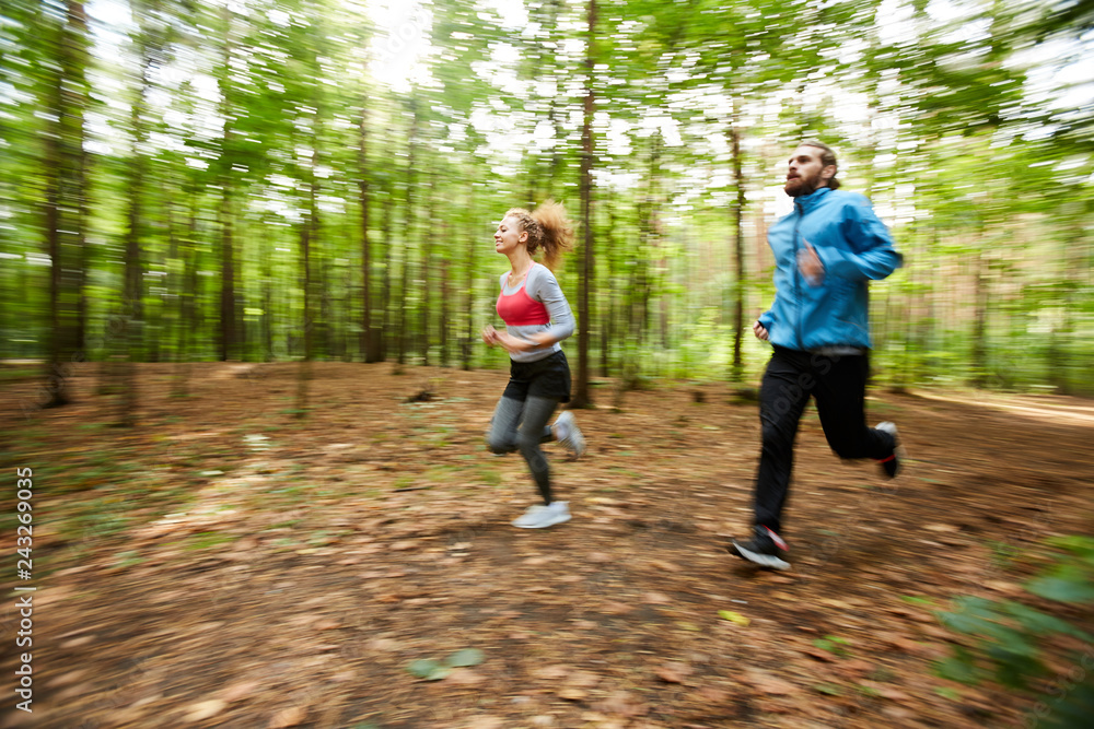 Active young couple in sportswear running down forest road on warm summer morning with blurry trees on background Active young couple in sportswear running down forest road on warm summer morning with blurry trees on background