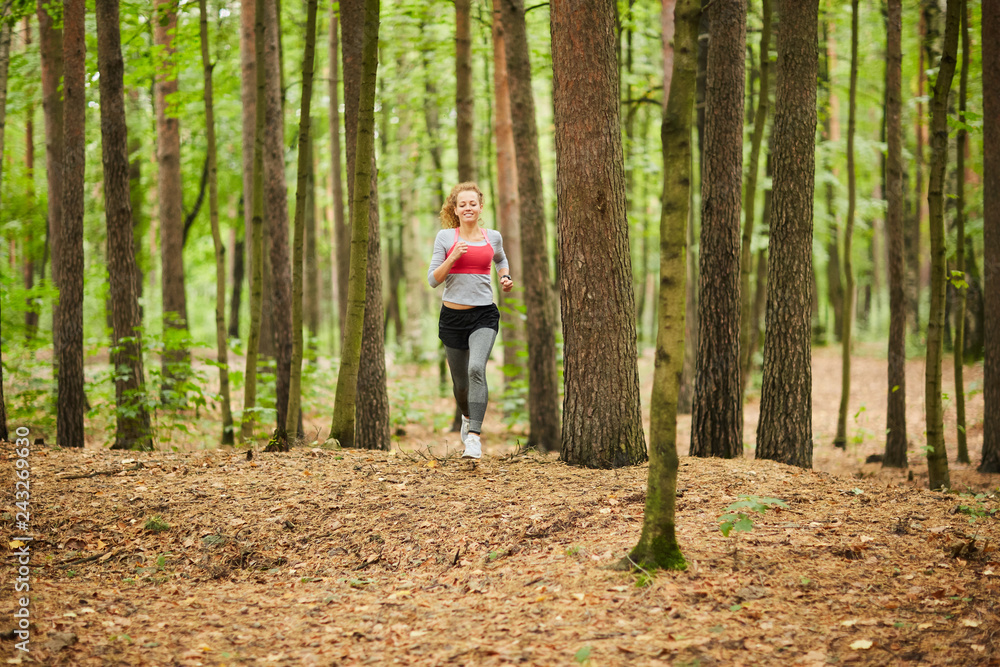 Obraz premium Young sportswoman running down forest path among trees while training on summer morning